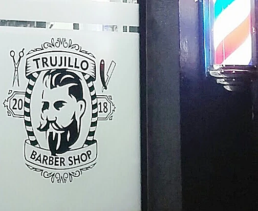 Puerta de vidrio de noche con el logo de Trujillo Barber Shop grabado, un barbudo con tijeras y navaja y el año 2018, junto a un poste de barbero encendido