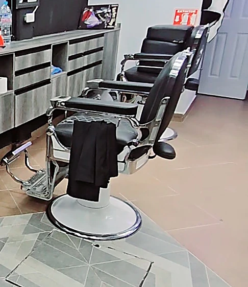 Dos sillones de barbero de cuero negro y cromo frente al mostrador gris, sobre el mosaico en espiga del piso